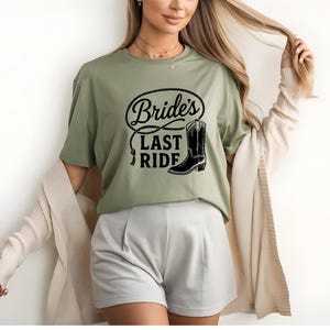 Könnte beinhalten: Salbeigrünes T-Shirt mit dem Text "Bride's Last Ride" in schwarzer Schrift und einer schwarzen Cowboy-Stiefel-Grafik. Das Design ist von einem Seil umgeben. Das Shirt ist ein Freizeitkleidungsstück.