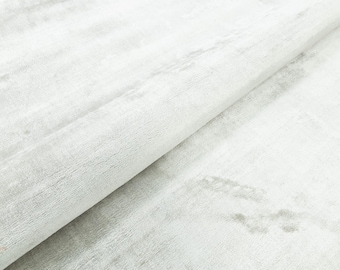 Puede incluir: Primer plano de una tela blanca con un aspecto texturizado y cepillado. El material presenta sutiles variaciones tonales, creando una estética suave y fluida. La tela está doblada, mostrando profundidad y dimensión.