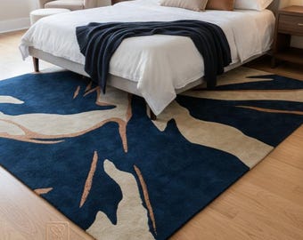 May include: A large, rectangular area rug with a modern design. The rug features a navy blue background with beige and copper-colored abstract shapes. The rug is placed in a bedroom setting, under a bed with white bedding.