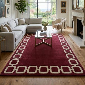 May include: A rectangular area rug featuring a deep burgundy red base colour with a contrasting cream-coloured border pattern consisting of a continuous chain of rounded square links. The rug is positioned on a light-toned wooden floor.