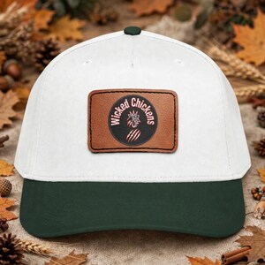 May include: A white baseball cap with a dark green brim and a dark green button on top. Centered on the front is a rectangular brown leather patch featuring a black circular emblem with the text Wicked Chickens and a rooster graphic.