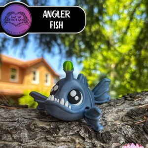 May include: A blue Angler Fish figurine with large white teeth, big black eyes, and a green lure. The figurine is resting on a piece of wood, with the text "Angler Fish" in a black and white banner.