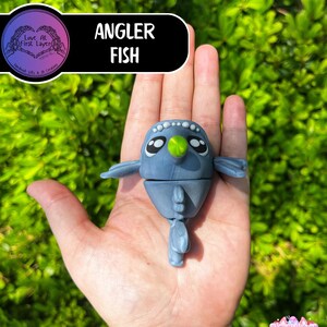 May include: A blue Angler Fish figurine with a green lure, held in a hand against a green leafy background. The figurine has large eyes and small fins. The text "Angler Fish" is displayed on a black banner.
