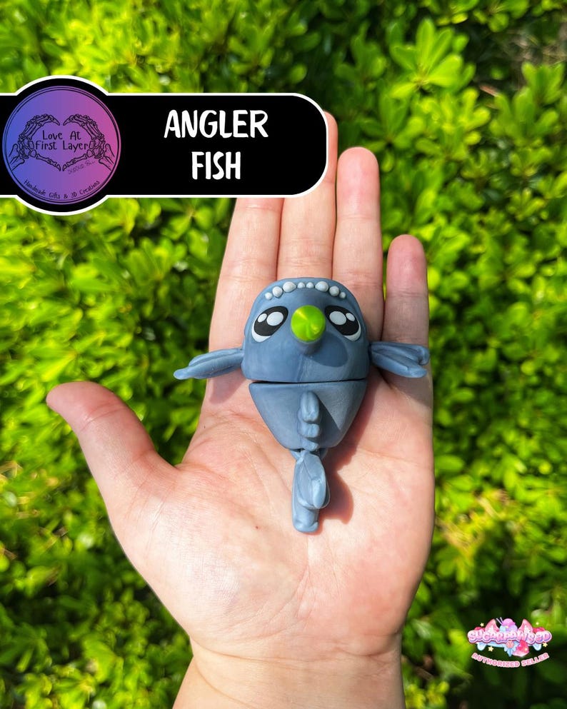 May include: A blue Angler Fish figurine with a green lure, held in a hand against a green leafy background. The figurine has large eyes and small fins. The text "Angler Fish" is displayed on a black banner.