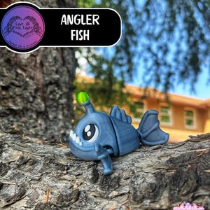May include: A blue angler fish figurine with a glowing green lure. The figurine has large eyes, sharp teeth, and fins. The text "Angler Fish" is displayed on a black background. The figurine is resting on a tree branch.