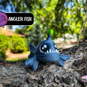 May include: A blue angler fish figurine with large eyes, white teeth, and a green light on its head. The figurine is resting on a textured, brown surface. The text "Angler Fish" is displayed above the figurine.