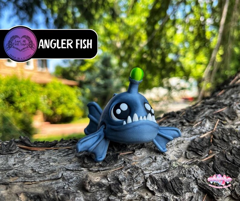 May include: A blue angler fish figurine with large eyes, white teeth, and a green light on its head. The figurine is resting on a textured, brown surface. The text "Angler Fish" is displayed above the figurine.