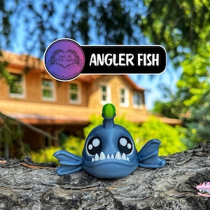 May include: A blue angler fish figurine with large, expressive eyes and a green lure. The figurine is set against a blurred background of a house and greenery. The text "ANGLER FISH" is displayed on a black banner.