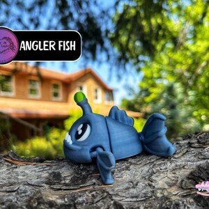 May include: A blue Angler Fish figurine with a green light on its head, resting on a textured surface. The figurine has large white eyes and fins. The text "Angler Fish" is displayed on a black banner.