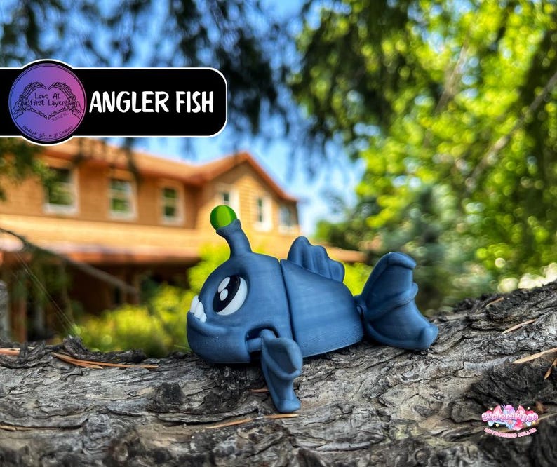 May include: A blue Angler Fish figurine with a green light on its head, resting on a textured surface. The figurine has large white eyes and fins. The text "Angler Fish" is displayed on a black banner.