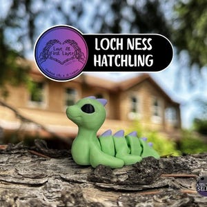 May include: A green Loch Ness Hatchling figurine with purple accents, resting on a textured surface. The figurine has black eyes and a stylized design. The image includes a logo with the text "Love At First Layer" and "Loch Ness Hatchling".