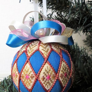 May include: A blue and gold Christmas ornament with a diamond pattern and pink accents. The ornament is decorated with a white, pink, and blue ribbon.