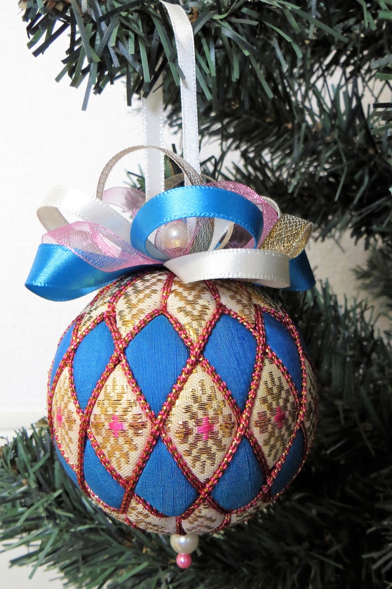 May include: A blue and gold Christmas ornament with a diamond pattern and pink accents. The ornament is decorated with a white, pink, and blue ribbon.