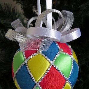 May include: A handmade Christmas ornament with a quilted design. The ornament is made of red, blue, green, and yellow fabric with silver trim. It is decorated with white beads and has a white ribbon and silver mesh bow.