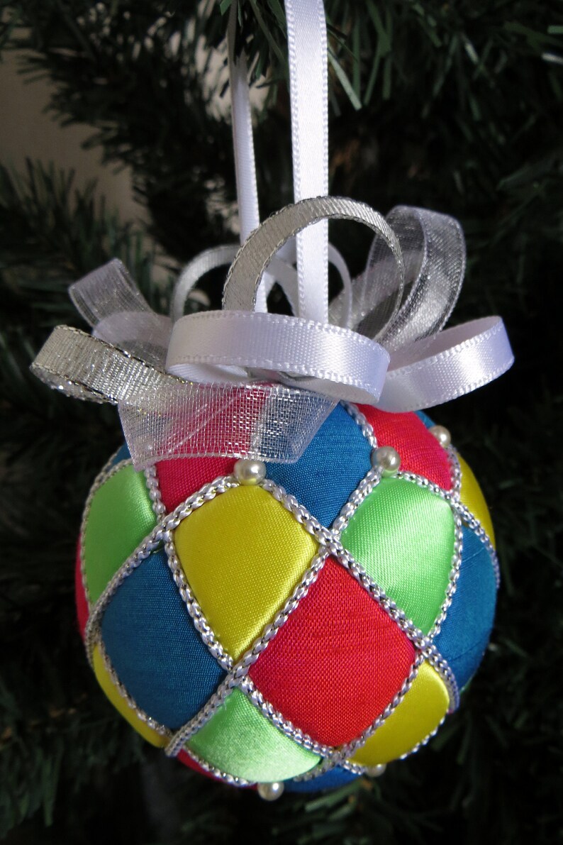 May include: A handmade Christmas ornament with a quilted design. The ornament is made of red, blue, green, and yellow fabric with silver trim. It is decorated with white beads and has a white ribbon and silver mesh bow.