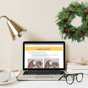 May include: A laptop computer displaying a webpage with instructions on how to make a Christmas wreath. The webpage shows a step-by-step guide with illustrations. The laptop is on a white desk with a cup of coffee, a gold lamp, a stack of books, a small white bowl, and a green Christmas wreath hanging on the wall.