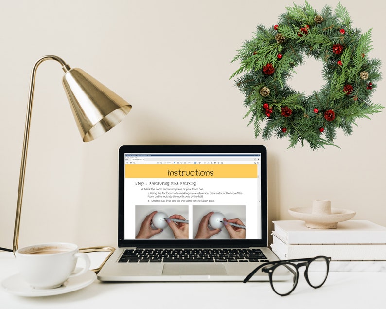 May include: A laptop computer displaying a webpage with instructions on how to make a Christmas wreath. The webpage shows a step-by-step guide with illustrations. The laptop is on a white desk with a cup of coffee, a gold lamp, a stack of books, a small white bowl, and a green Christmas wreath hanging on the wall.