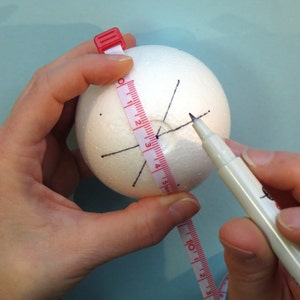 May include: A white styrofoam ball with black lines drawn on it. A person is using a measuring tape and a black marker to mark the ball.