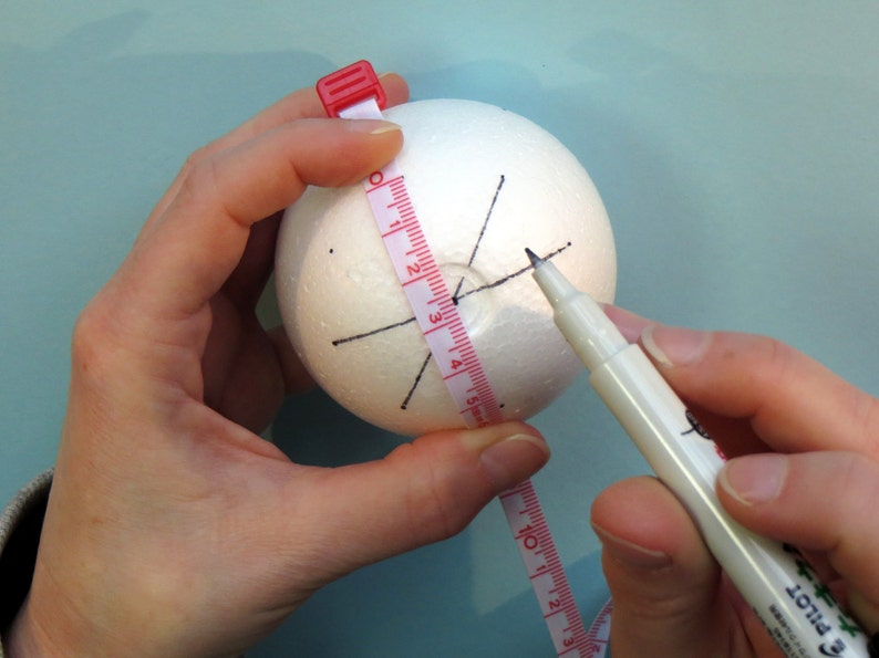 May include: A white styrofoam ball with black lines drawn on it. A person is using a measuring tape and a black marker to mark the ball.