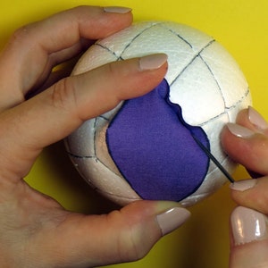 May include: A white styrofoam ball with a grid pattern drawn on it. A purple fabric patch is being sewn onto the ball with a needle and thread.