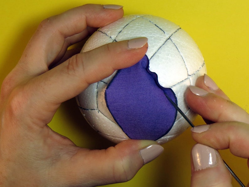 May include: A white styrofoam ball with a grid pattern drawn on it. A purple fabric patch is being sewn onto the ball with a needle and thread.