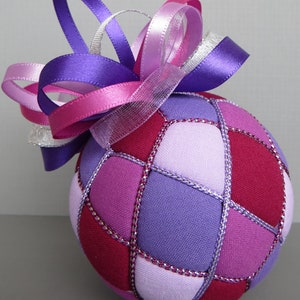 May include: A purple, pink, and white fabric Christmas ornament with a large bow. The ornament is made of fabric squares sewn together with silver thread.