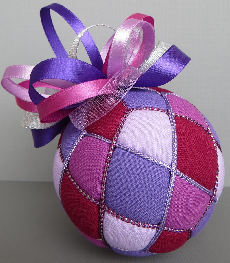 May include: A purple, pink, and white fabric Christmas ornament with a large bow. The ornament is made of fabric squares sewn together with silver thread.