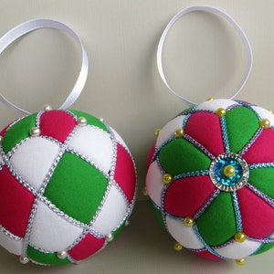 May include: Two handmade Christmas ornaments, one with a green, white, and pink diamond pattern and the other with a green, white, and pink flower pattern. Both ornaments are decorated with white and gold beads.
