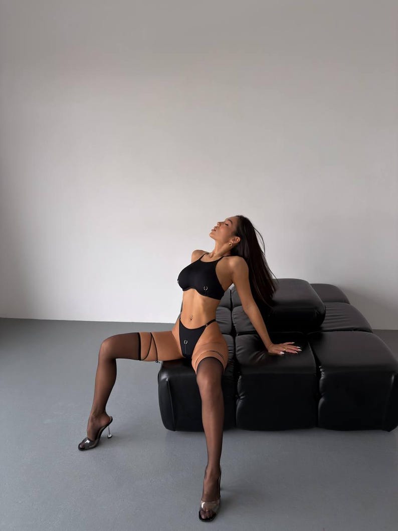 May include: Black lingerie set featuring a bralette and high-waisted bottoms, accented with silver rings. The model is wearing sheer black stockings with garter straps and clear high heels. The background is a minimalist white wall and a black leather couch.
