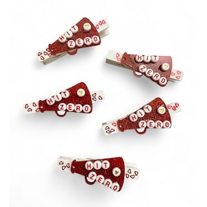 Può includere: Cinque fermagli per capelli rossi con glitter a forma di megafono con lettere bianche che compongono la scritta "HIT ZERO" e piccoli cuori bianchi.