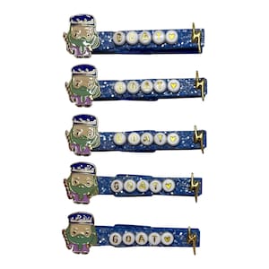 May include: Five blue glitter hair clips with a gold lightning bolt charm and a cartoon character with a beard and a crown. Each clip has the word "GOAT" spelled out in gold letters on white beads.