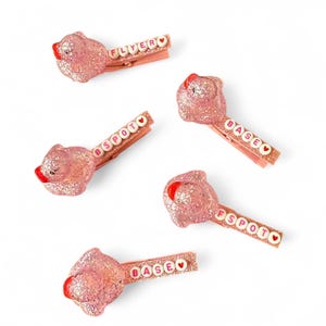 Puede incluir: Cinco pinzas para el pelo de purpurina rosa con un pequeño encanto de cerdo rosa en la parte superior. Cada pinza tiene una palabra diferente escrita en letras blancas con un corazón rosa al final. Las palabras son "FLYER", "BSPOT", "BASE", "FSPOT" y "BASE".