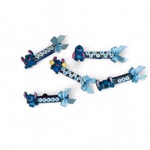 May include: Five blue hair clips featuring the character Stitch from Lilo & Stitch. Each clip has a different word spelled out in white letters, including "Aloha", "Ohana", "Boo Boo", "Achiba", and "Tay". Each clip has a blue bow.