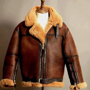 May include: A brown leather jacket with a shearling collar and lining. The jacket features a zip-up front, a belt at the waist, and zippered sleeves. The jacket is displayed on a mannequin.