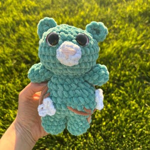 Könnte beinhalten: Ein handgefertigter, türkisfarbener Häkelbär mit großen schwarzen Augen und einer weißen Schnauze. Der Bär hat kleine Ohren und Arme und hält ein kleines braun-weißes Objekt. Der Bär ist aus weichem, strukturiertem Garn gefertigt.