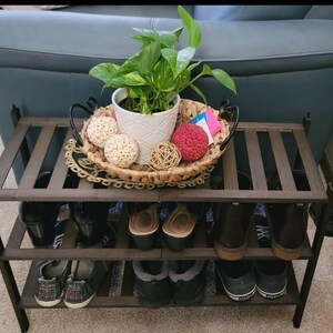 Puede incluir: Un zapatero de madera marrón oscuro con tres estantes. El estante está lleno de zapatos, incluyendo botas, zapatillas y sandalias. Una planta en maceta con hojas verdes y bolas decorativas blancas se encuentra en la parte superior del estante.