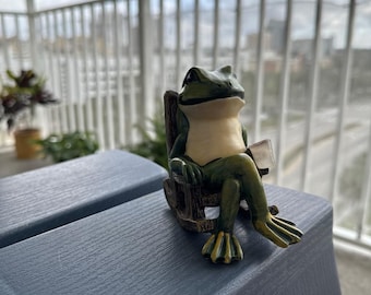 Könnte beinhalten: Eine grüne Froschfigur, die auf einem Schaukelstuhl sitzt und eine kleine Tasse hält. Der Frosch sitzt auf einem blauen Plastikhocker.