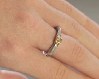Könnte beinhalten: Ein silberner Ring mit einer strukturierten Band und einem kleinen, quadratischen Diamanten, der in einer goldenen Fassung sitzt.