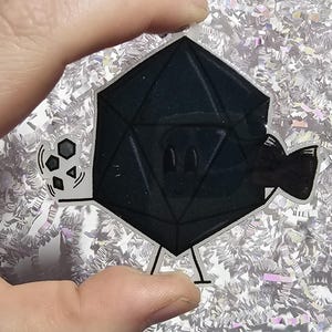 May include: A black, twenty-sided die character sticker with a white outline. The die has a simple face with two oval eyes and a small smile. One arm holds a smaller die, while the other arm is bent. The sticker has a clear, transparent border.