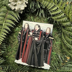 May include: Enamel pin depicting three figures in black and red capes, set against a snowy backdrop. The figures have long hair and are in a dramatic pose. The pin is surrounded by green foliage.