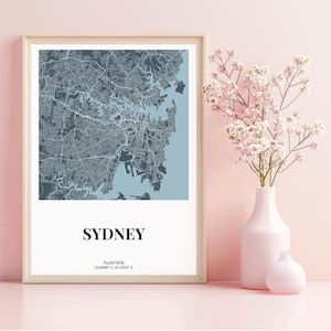 May include: Framed map print of Sydney, Australia, in shades of gray and blue. The word "SYDNEY" is printed in a bold, black font at the bottom. A pink vase with flowers is to the right.