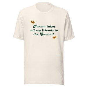 May include: Cream-colored t-shirt with the text "Karma takes all my friends to the Summit" in green script. The shirt features two sets of gold starburst graphics, one above and one below the text.