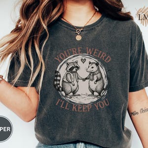 May include: Dark gray t-shirt featuring a raccoon and opossum holding hands inside a circle with the text "You're Weird, I'll Keep You." The graphic is in shades of gray and brown. The shirt has a vintage, worn look.