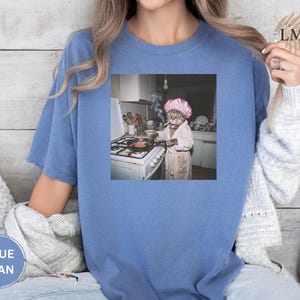 May include: Blue jean colored t-shirt with a square graphic. The graphic depicts a cat wearing a pink shower cap and robe, cooking in a kitchen. The cat is smoking a cigarette while cooking.