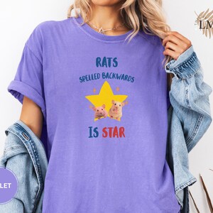 May include: Violet-colored t-shirt with the text "RATS SPELLED BACKWARDS IS STAR" in blue and red. A yellow star graphic with two cartoon rats is centered on the shirt. The shirt has rolled-up sleeves.