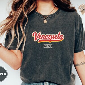 Pu&ograve; includere: T-shirt grigio scuro con la scritta "Venezuela" in rosso con contorno giallo e l'anno "2026". La maglietta &egrave; di stile casual.
