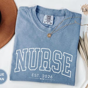 May include: A blue jean Comfort Colors t-shirt with the word "NURSE" in white block letters. Below the word "NURSE" is the text "EST. 2026" and three small hearts. A gold necklace and a brown hat are also in the image.