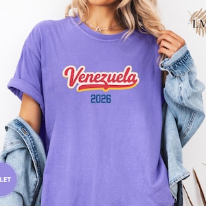 Pu&ograve; includere: T-shirt viola con la scritta "Venezuela" in corsivo rosso e bianco con contorno giallo. Sotto la parola c'&egrave; il numero "2026" in blu. Le maniche della maglietta sono arrotolate.