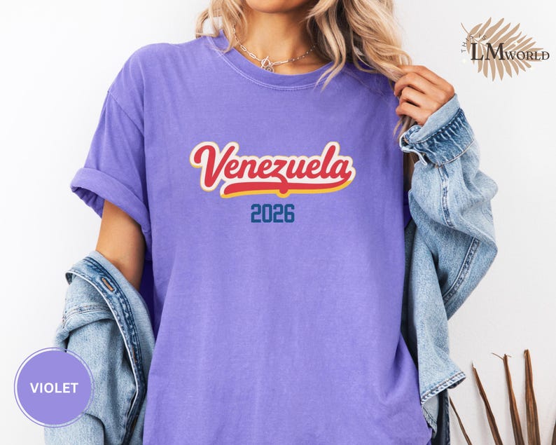 Pu&ograve; includere: T-shirt viola con la scritta "Venezuela" in corsivo rosso e bianco con contorno giallo. Sotto la parola c'&egrave; il numero "2026" in blu. Le maniche della maglietta sono arrotolate.