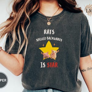 May include: Dark gray t-shirt with the text "RATS SPELLED BACKWARDS IS STAR" in white and red. The design includes a yellow star and two cartoon rats. The shirt is a comfortable, casual style.
